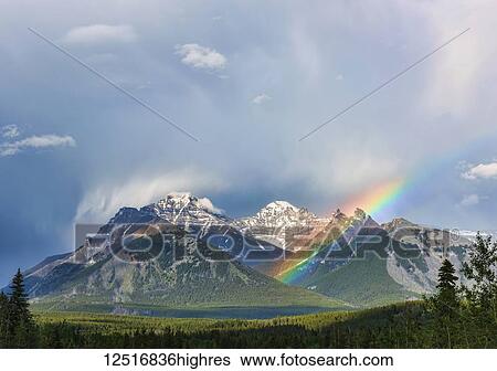 Stock Photograph - A rainbow emerges between rugged mountain peaks during a heavy rain storm; Banff, Alberta, Canada. Fotosearch
