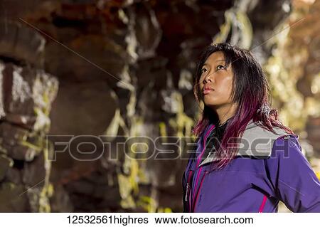 Stock Photograph - A side profile close-up of an asian female hiker looking upwards at the cave walls in a lava tube cave; Iceland. Fotosearch