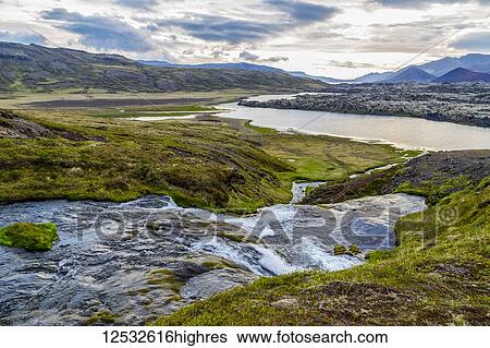 Stock Photograph - A small river runs through a valley in Western Iceland towards the large river below and the distant richly colored volcanic peaks in the distance; Iceland. Fotosearch