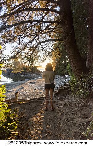 Stock Photograph - A teenage girl stands on the sand of Tonquin Beach at sunset looking out to the water; Tofino, British Columbia, Canada. Fotosearch