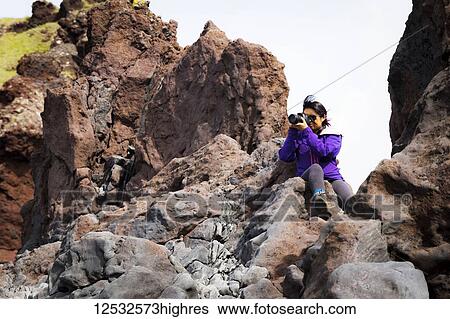 Stock Photograph - A tourist sits on the top of a rock formation to take a higher photo of the landscape; Iceland. Fotosearch