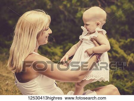 Stock Photograph - A vintage style image of a beautiful young mother with long blonde hair enjoying quality time with her cute baby daughter sitting on the grass in a city park on a summer day; Edmonton, Alberta, Canada. Fotosearch