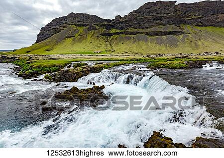 Stock Photograph - A wide angle of the river cascading over the rocks in front of a volcanic mountain and farmland; Iceland. Fotosearch