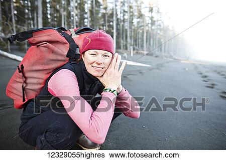 Stock Photograph - A woman crouching on the beach carrying a backpack, Kenai Peninsula outer coast; Alaska, United States of America. Fotosearch
