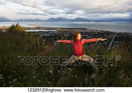 Stock Photograph - A woman doing yoga in a meadow with a view of Homer Spit, Kachemak Bay and the Kenai Mountains in the background; Homer, Alaska, United States of America. Fotosearch