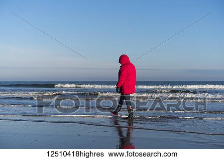 Stock Photograph - A woman in a red coat and rubber boots walks in the shallow water along the shore with the blue sky and ocean; Bamburgh, Northumberland, England. Fotosearch