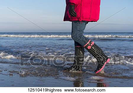 Stock Photograph - A woman in a red coat and rubber boots walks in the shallow water along the shore with the blue sky and ocean; Bamburgh, Northumberland, England. Fotosearch