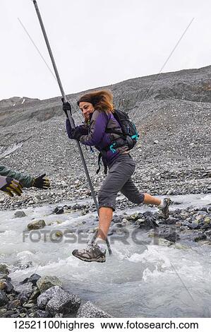 Stock Photograph - A woman jumps over a swift glacial stream with the aid of a pole near Gulkana Glacier; Alaska, United States of America. Fotosearch