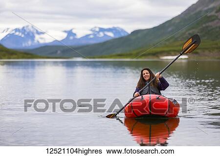 Stock Photograph - A woman paddles a packraft across Landmark Gap Lake, off the Denali highway, with the Alaska Range in the distance; Alaska, United States of America. Fotosearch
