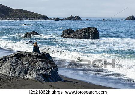Stock Photograph - A woman sits on a rock on the beach watching the waves coming ashore along the rocky California coast; Crescent City, California, United States of America. Fotosearch