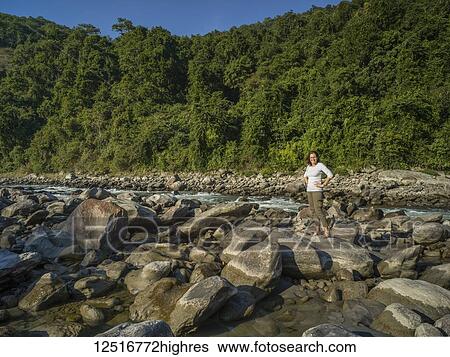 Stock Photograph - A woman stands on a large rock along the shoreline of a river with a lush forest in the background; West Bengal, India. Fotosearch