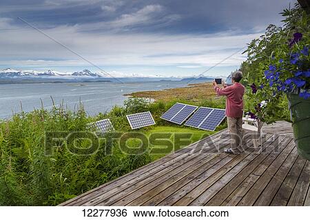 Stock Photograph - A woman takes a photo of the Chilkat Mountain Range with solar panels in the foreground, Southeast Alaska, Summer.. Fotosearch