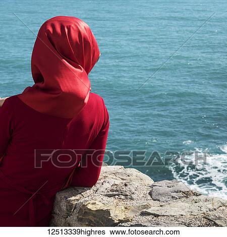 Stock Photograph - A woman wearing a red headscarf and red dress stands at a stone wall looking out at the blue water; Budva, Budva Municipality, Monte. Fotosearch