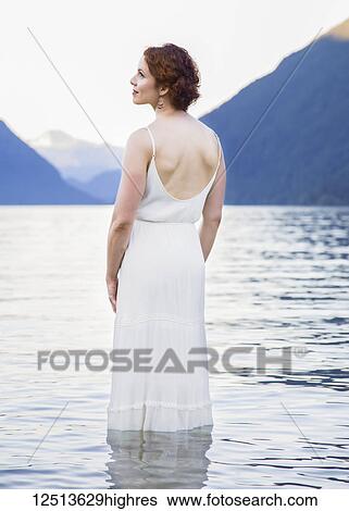 Stock Photograph - A woman with red hair and a white dress stands in the tranquil water of Alouette Lake on a summer evening; British Columbia, Canada. Fotosearch