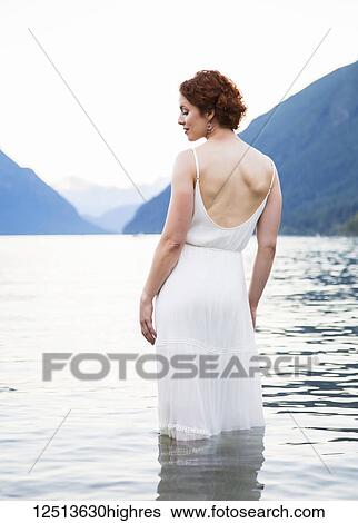 Stock Photograph - A woman with red hair and a white dress stands in the tranquil water of Alouette Lake on a summer evening; British Columbia, Canada. Fotosearch