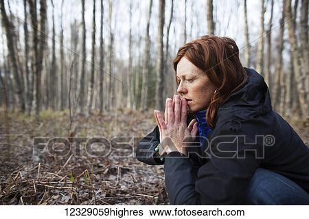 Stock Photograph - A woman with red hair crouches on a forest floor with her hands pressed together; Alaska, United States of America. Fotosearch