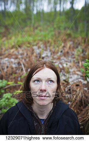 Stock Photograph - A woman with red hair looks up to the sky with a forest in the background; Alaska, United States of America. Fotosearch