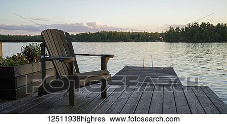 A wooden Adirondack chair sits on a dock on a tranquil lake at sunset; Lake of the Woods, Ontario, Canada View Large Photo Image Stock Photograph - A wooden Adirondack chair sits on a dock on a tranquil lake at sunset; Lake of the Woods, Ontario, Canada. Fotosearch