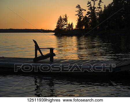 A wooden Adirondack chair sits on a dock on a tranquil lake at sunset; Lake of the Woods, Ontario, Canada View Large Photo Image Stock Photograph - A wooden Adirondack chair sits on a dock on a tranquil lake at sunset; Lake of the Woods, Ontario, Canada. Fotosearch