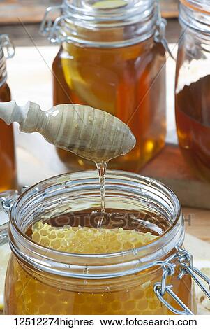 Stock Photograph - A wooden dipper dripping above a jar of honey with honeycomb. Fotosearch