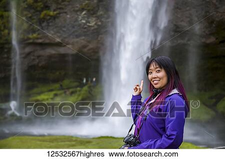 Stock Photograph - A young asian female hiker poses for a portrait at a waterfall; Iceland. Fotosearch