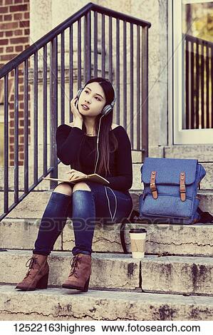 A young Chinese female university student sits on the steps listening to music using headphones and looks away while daydreaming; Edmonton, Alberta, Canada View Large Photo Image Stock Photograph - A young Chinese female university student sits on the steps listening to music using headphones and looks away while daydreaming; Edmonton, Alberta, Canada. Fotosearch