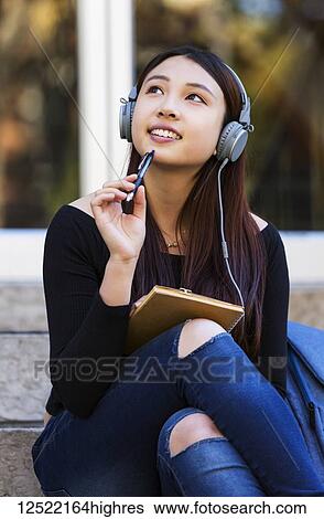 Stock Photograph - A young Chinese female university student sits on the steps listening to music using headphones and looks up while daydreaming and holding a pen top to her chin; Edmonton, Alberta, Canada. Fotosearch