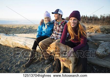 Stock Photograph - A young couple and a friend with a dog sit on a piece of driftwood on a beach looking out to the ocean at sunset; Anchorage, Alaska, United States of America. Fotosearch