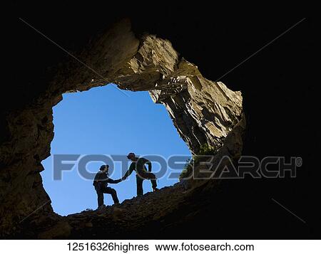 Stock Photograph - A young couple climbing in the mouth of a cave, holding hands for support; Fernie, British Columbia, Canada. Fotosearch