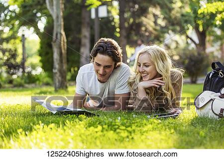 Stock Photograph - A young couple lays down studying on the grass on the university campus looking through a textbook and using a tablet; Edmonton, Alberta, Canada. Fotosearch