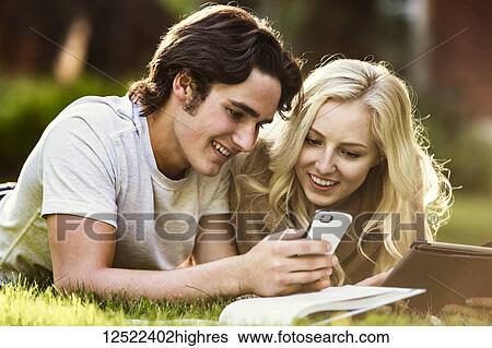 Stock Photograph - A young couple lays down studying outdoors on the grass on the university campus with a textbook and tablet and checking social media on a smart phone; Edmonton, Alberta, Canada. Fotosearch