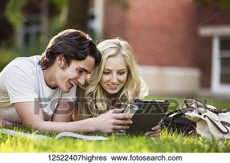 Stock Photograph - A young couple lays down studying outdoors on the grass on the university campus with a textbook and tablet and checking social media on a smart phone; Edmonton, Alberta, Canada. Fotosearch