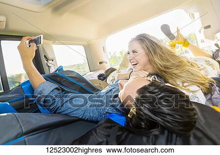 Stock Photograph - A young couple lays in the back of their vehicle during a road trip talking and laughing together and taking a self-portrait with their cell phone; Edmonton, Alberta, Canada. Fotosearch