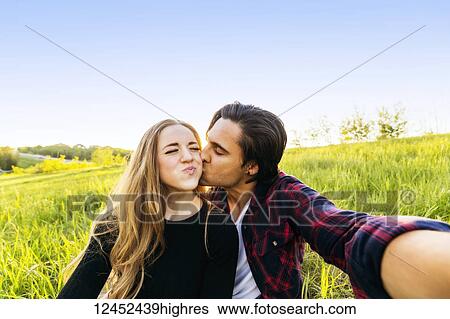 Stock Photograph - A young couple sits in a field taking a self-portrait while the young man kisses the young woman on the cheek; Edmonton, Alberta, Canada. Fotosearch