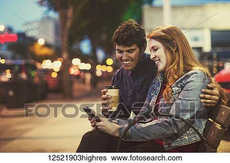 Stock Photograph - A young couple sits on a bench along a popular street at dusk looking at a smart phone; Edmonton, Alberta, Canada. Fotosearch