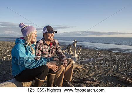 Stock Photograph - A young couple sits on a piece of driftwood on a beach looking out to the ocean at sunset; Anchorage, Alaska, United States of America. Fotosearch