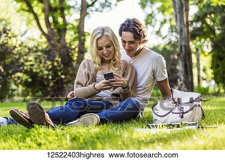 Stock Photograph - A young couple sits studying on the grass on the university campus with a textbook and tablet while using a smart phone; Edmonton, Alberta, Canada. Fotosearch
