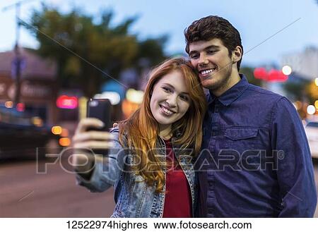 Stock Photograph - A young couple stand beside a city street at dusk taking a self- portrait with a smart phone; Edmonton, Alberta, Canada. Fotosearch