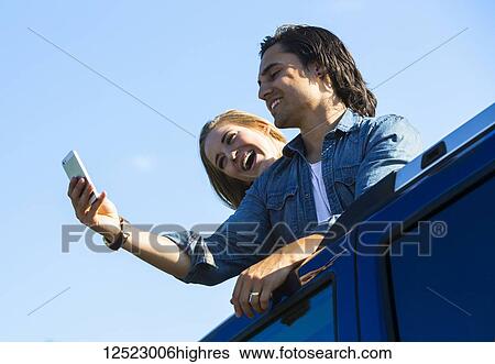 Stock Photograph - A young couple stand up through the sunroof of their vehicle and take a self-portrait with their cell phone; Edmonton, Alberta, Canada. Fotosearch