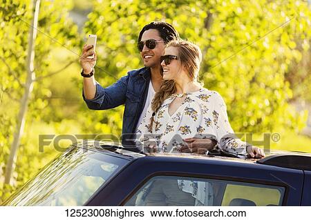Stock Photograph - A young couple stand up through the sunroof of their vehicle and take a self-portrait with their cell phone; Edmonton, Alberta, Canada. Fotosearch