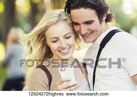 Stock Photograph - A young couple standing together and checking social media on a smart phone while walking through a university campus; Edmonton, Alberta, Canada. Fotosearch