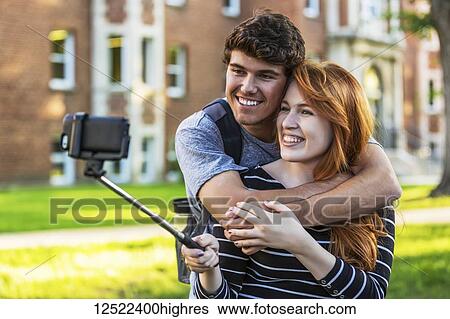 Stock Photograph - A young couple standing together in a university campus and taking a selfie with a smart phone on a selfie stick; Edmonton, Alberta, Canada. Fotosearch