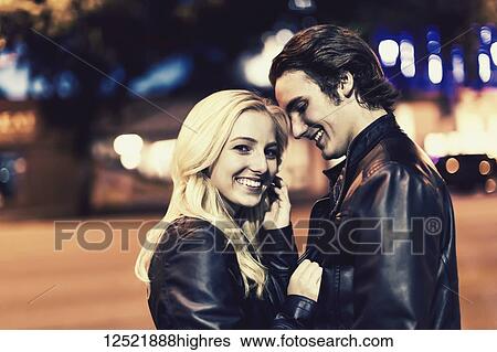 Stock Photograph - A young couple stands close on the sidewalk beside a city street at dusk and she poses for the camera; Edmonton, Alberta, Canada. Fotosearch