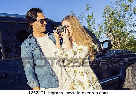 Stock Photograph - A young couple stands outside their vehicle on a road trip and the young woman takes a photograph with her camera; Edmonton, Alberta, Canada. Fotosearch