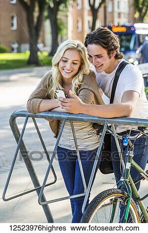Stock Photograph - A young couple stands together at a bike rack on the university campus looking at a smart phone; Edmonton, Alberta, Canada. Fotosearch