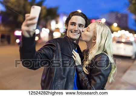 A young couple take a self-portrait with a smart phone as the female kisses the male on the cheek along a street at dusk; Edmonton, Alberta, Canada View Large Photo Image Stock Photograph - A young couple take a self-portrait with a smart phone as the female kisses the male on the cheek along a street at dusk; Edmonton, Alberta, Canada. Fotosearch