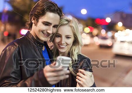 Stock Photograph - A young couple use a smart phone along a street at dusk; Edmonton, Alberta, Canada. Fotosearch