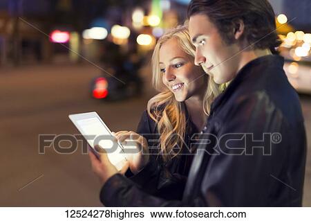 Stock Photograph - A young couple use a tablet along a street at dusk with the glowing screen illuminating their faces; Edmonton, Alberta, Canada. Fotosearch