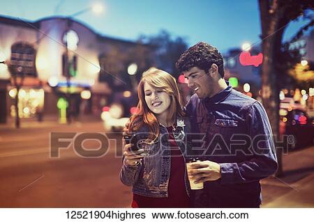 A young couple walking down a sidewalk in a popular area at dusk while looking at a smart phone; Edmonton, Alberta, Canada View Large Photo Image Stock Photograph - A young couple walking down a sidewalk in a popular area at dusk while looking at a smart phone; Edmonton, Alberta, Canada. Fotosearch