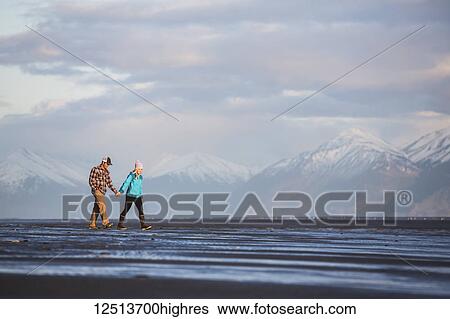 Stock Photograph - A young couple walking on a beach and holding hands with a mountain range in the distance; Anchorage, Alaska, United States of America. Fotosearch